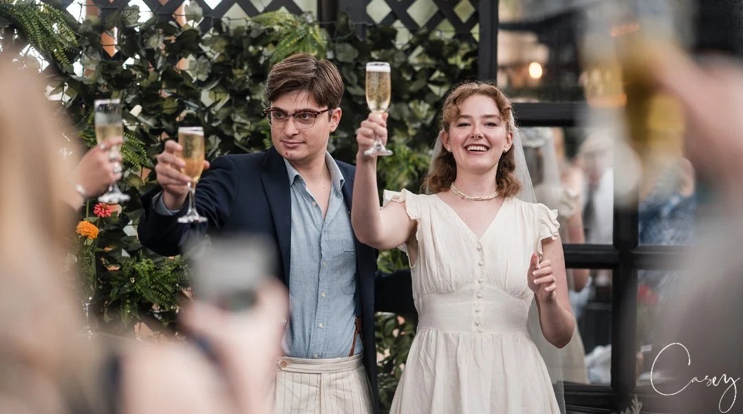 A couple raising champagne glasses in a toast at a wedding celebration, with greenery behind them